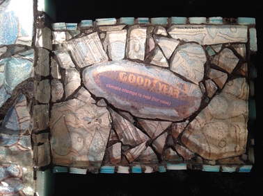 glass mosaic on wood / detail / photo of the Goodyear blimp under glass / text riding on blimp: climate change is real ( for now)