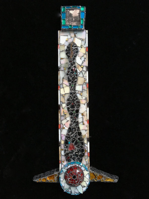 glass mosaic totem on wood with photograph from Portugal or Spain on the top --- an image of a goddess. The piece has a black snakelike form going down the length of the piece and on the bottom, there is a red round shape, kind of like an eye or an orb surrounded by white triangles in a white background. The circle is ringed with turquoise glass and there are amber colored glass triangles like flippers on the bottom.